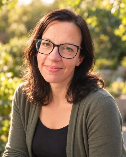 Daniela Hirzer mit Brille und olivgrüner Strickjacke im sonnigen Garten